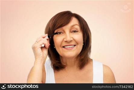 beauty, make up and old people concept - smiling senior woman applying mascara to eyelashes over beige background. smiling senior woman applying mascara to eyelashes