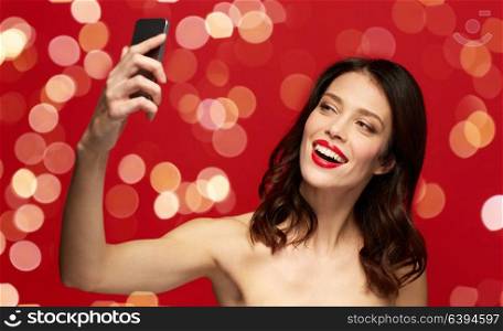 beauty, make up and people concept - happy smiling young woman with red lipstick taking selfie by smartphone over red background with lights. beautiful woman taking selfie by smartphone