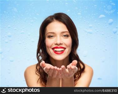 beauty, make up and skin moisturizing concept - happy smiling young woman with red lipstick holding something imaginary on palms over water drops on blue glass background . beautiful smiling young woman with red lipstick. beautiful smiling young woman with red lipstick