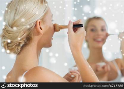 beauty, make up, cosmetics, morning and people concept - close up of smiling young woman makeup brush and powder foundation looking to mirror at home bathroom over snow