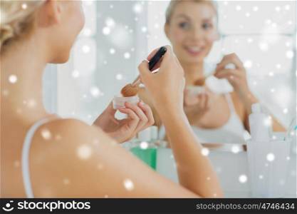 beauty, make up, cosmetics, morning and people concept - close up of young woman makeup brush and powder foundation looking to mirror at home bathroom over snow