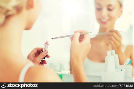 beauty, make up, cosmetics, morning and people concept - close up of smiling young woman with lipstick and make up brush at home bathroom mirror. woman with lipstick and make up brush at bathroom