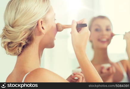 beauty, make up, cosmetics, morning and people concept - close up of smiling young woman makeup brush and powder foundation looking to mirror at home bathroom. woman with makeup brush and powder at bathroom. woman with makeup brush and powder at bathroom