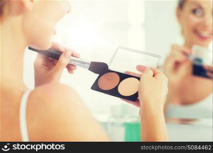beauty, make up, cosmetics, morning and people concept - close up of happy young woman with makeup brush and contouring and highlighting palette looking to mirror at home bathroom. woman with makeup brush and foundation at bathroom. woman with makeup brush and foundation at bathroom