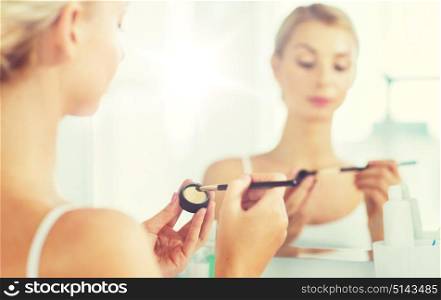 beauty, make up, cosmetics, morning and people concept - close up of young woman applying eyeshade with makeup brush and looking to mirror at home bathroom. woman with makeup brush and eyeshade at bathroom
