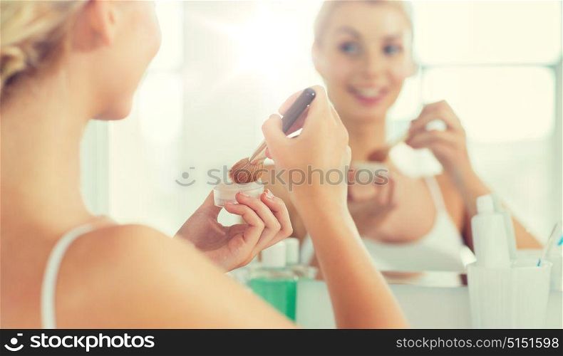 beauty, make up, cosmetics, morning and people concept - close up of young woman makeup brush and powder foundation looking to mirror at home bathroom. woman with makeup brush and powder at bathroom