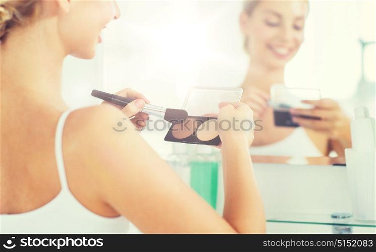 beauty, make up, cosmetics, morning and people concept - close up of happy young woman with makeup brush and contouring and highlighting palette looking to mirror at home bathroom. woman with makeup brush and foundation at bathroom