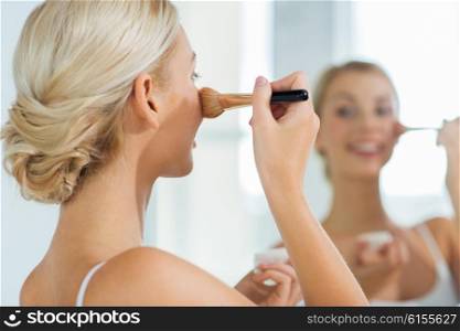beauty, make up, cosmetics, morning and people concept - close up of smiling young woman makeup brush and powder foundation looking to mirror at home bathroom