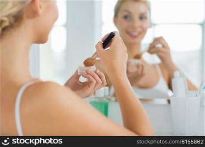 beauty, make up, cosmetics, morning and people concept - close up of young woman makeup brush and powder foundation looking to mirror at home bathroom