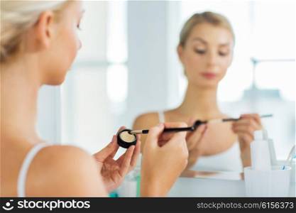 beauty, make up, cosmetics, morning and people concept - close up of young woman applying eyeshade with makeup brush and looking to mirror at home bathroom