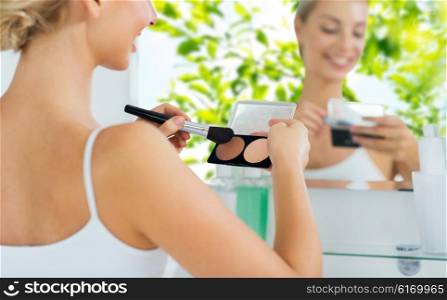 beauty, make up, cosmetics, morning and people concept - close up of happy young woman with makeup brush and contouring highlighting palette at bathroom over green natural background
