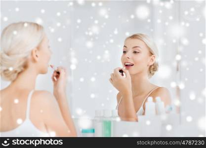 beauty, make up, cosmetics, morning and people concept - smiling young woman coloring her lips with lipstick looking to mirror at home bathroom over snow