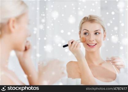beauty, make up, cosmetics, morning and people concept - smiling young woman makeup brush and powder foundation looking to mirror at home bathroom over snow