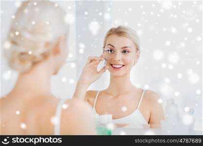 beauty, make up, cosmetics, morning and people concept - smiling young woman with curler curling eyelashes and looking to mirror at home bathroom over snow