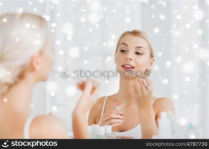beauty, make up, cosmetics, morning and people concept - smiling young woman with lipstick and applicator coloring her lips and looking to mirror at home bathroom over snow
