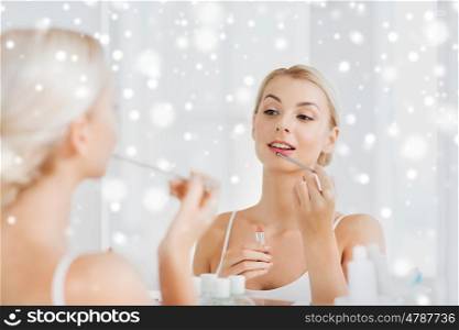 beauty, make up, cosmetics, morning and people concept - smiling young woman with lipstick and applicator coloring her lips and looking to mirror at home bathroom over snow