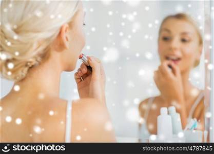 beauty, make up, cosmetics, morning and people concept - smiling young woman applying lipstick and looking to mirror at home bathroom over snow