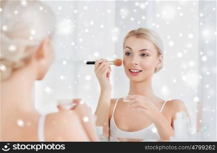 beauty, make up, cosmetics, morning and people concept - smiling young woman makeup brush and powder foundation looking to mirror at home bathroom over snow