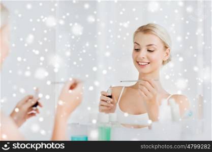 beauty, make up, cosmetics, morning and people concept - smiling young woman with lipstick and applicator at mirror at home bathroom over snow