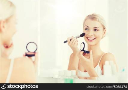 beauty, make up, cosmetics, morning and people concept - smiling young woman applying blush with makeup brush and looking to mirror at home bathroom. woman with makeup brush and blush at bathroom