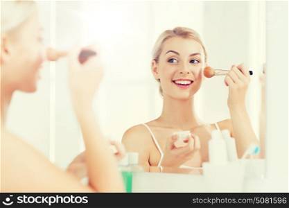 beauty, make up, cosmetics, morning and people concept - smiling young woman makeup brush and powder foundation looking to mirror at home bathroom. woman with makeup brush and powder at bathroom