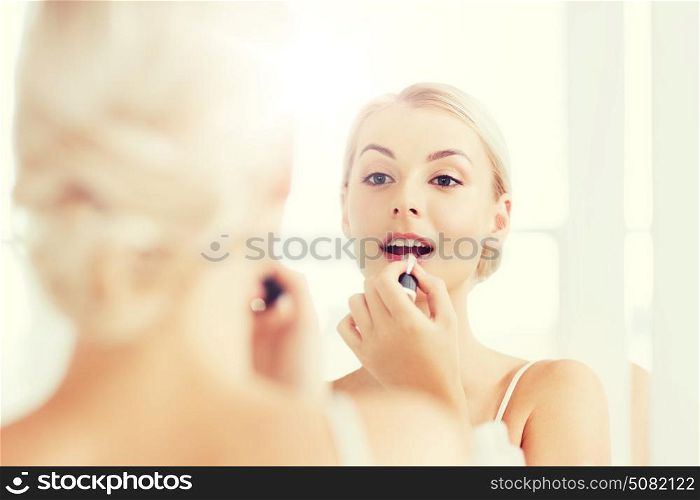 beauty, make up, cosmetics, morning and people concept - smiling young woman with lipstick applying makeup and looking to mirror at home bathroom. woman with lipstick applying make up at bathroom