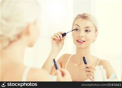 beauty, make up, cosmetics, morning and people concept - smiling young woman applying eye makeup with mascara and looking to mirror at home bathroom. woman with mascara applying make up at bathroom