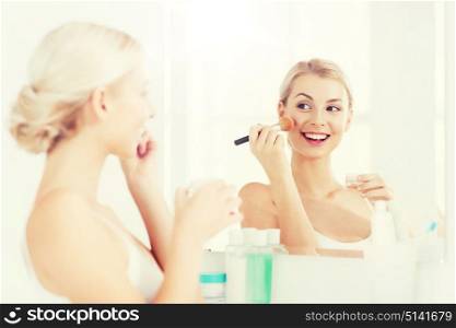 beauty, make up, cosmetics, morning and people concept - smiling young woman makeup brush and powder foundation looking to mirror at home bathroom. woman with makeup brush and powder at bathroom
