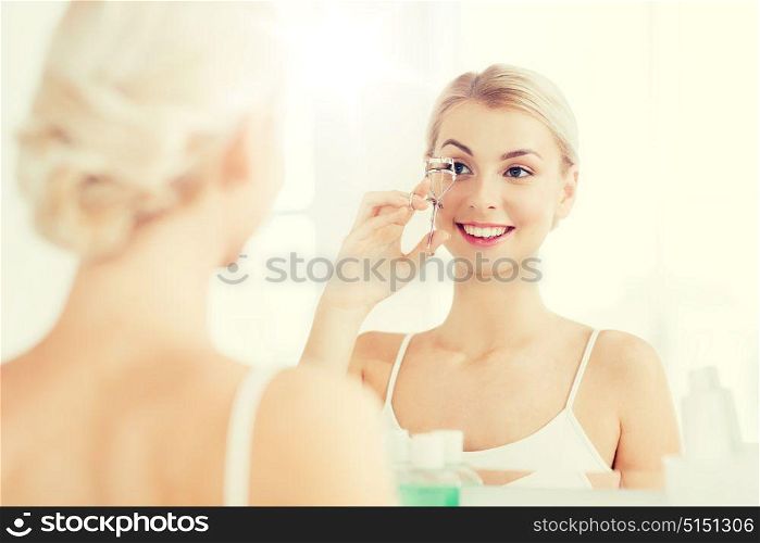 beauty, make up, cosmetics, morning and people concept - smiling young woman with curler curling eyelashes and looking to mirror at home bathroom. woman with curler curling eyelashes at bathroom