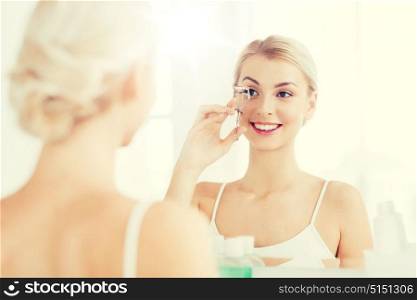 beauty, make up, cosmetics, morning and people concept - smiling young woman with curler curling eyelashes and looking to mirror at home bathroom. woman with curler curling eyelashes at bathroom
