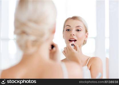 beauty, make up, cosmetics, morning and people concept - smiling young woman with lipstick applying makeup and looking to mirror at home bathroom