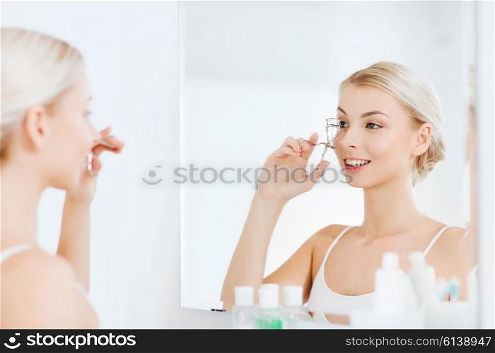 beauty, make up, cosmetics, morning and people concept - smiling young woman with curler curling eyelashes and looking to mirror at home bathroom