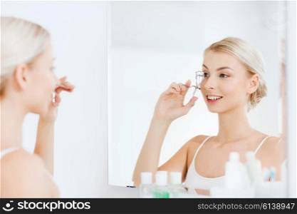 beauty, make up, cosmetics, morning and people concept - smiling young woman with curler curling eyelashes and looking to mirror at home bathroom