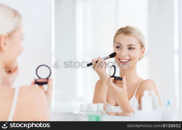 beauty, make up, cosmetics, morning and people concept - smiling young woman applying blush with makeup brush and looking to mirror at home bathroom