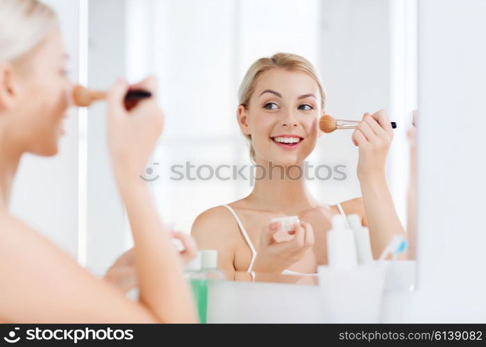 beauty, make up, cosmetics, morning and people concept - smiling young woman makeup brush and powder foundation looking to mirror at home bathroom