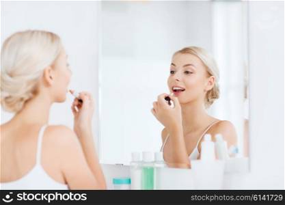 beauty, make up, cosmetics, morning and people concept - smiling young woman with lipstick applying makeup and looking to mirror at home bathroom