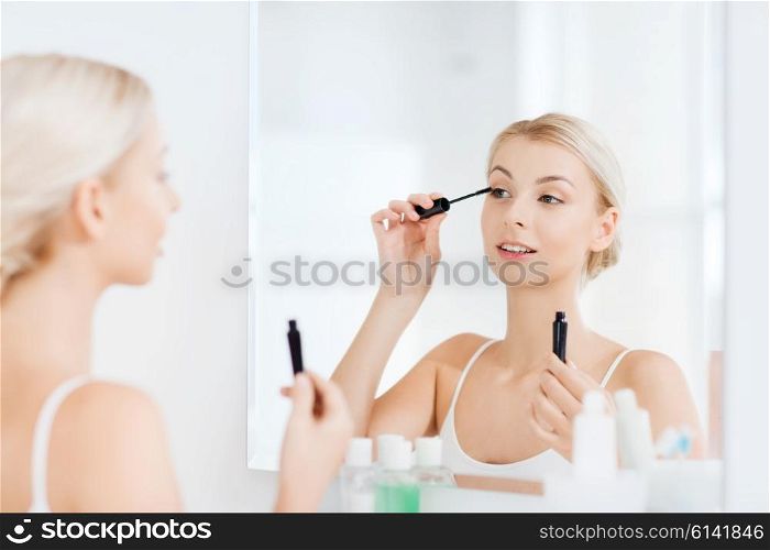 beauty, make up, cosmetics, morning and people concept - smiling young woman applying eye makeup with mascara and looking to mirror at home bathroom