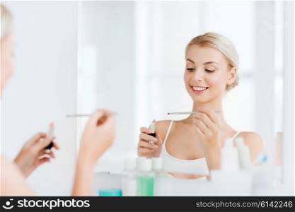beauty, make up, cosmetics, morning and people concept - smiling young woman with lipstick and applicator applying makeup and looking to mirror at home bathroom