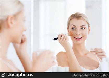 beauty, make up, cosmetics, morning and people concept - smiling young woman makeup brush and powder foundation looking to mirror at home bathroom