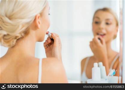 beauty, make up, cosmetics, morning and people concept - smiling young woman applying lipstick and looking to mirror at home bathroom