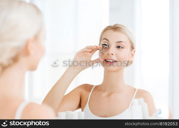 beauty, make up, cosmetics, morning and people concept - smiling young woman with curler curling eyelashes and looking to mirror at home bathroom