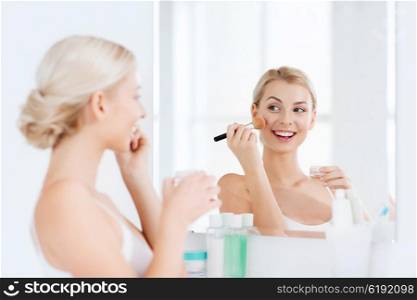 beauty, make up, cosmetics, morning and people concept - smiling young woman makeup brush and powder foundation looking to mirror at home bathroom