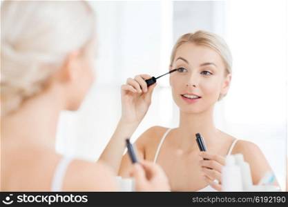 beauty, make up, cosmetics, morning and people concept - smiling young woman applying eye makeup with mascara and looking to mirror at home bathroom