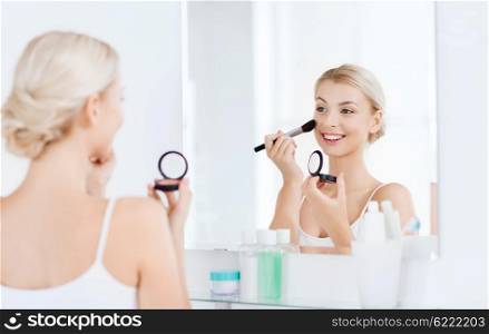 beauty, make up, cosmetics, morning and people concept - smiling young woman applying blush with makeup brush and looking to mirror at home bathroom