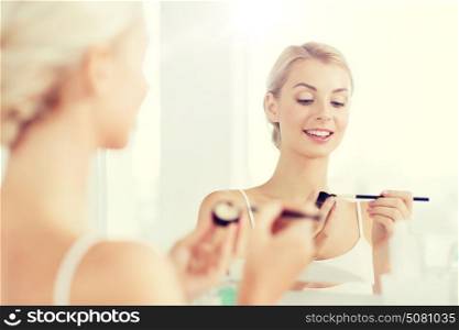 beauty, make up, cosmetics, morning and people concept - young woman applying eyeshade with makeup brush and looking to mirror at home bathroom. woman with makeup brush and eyeshade at bathroom