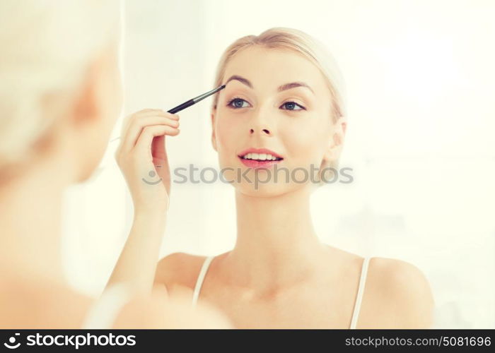 beauty, make up, cosmetics, morning and people concept - young woman applying makeup drawing eyebrow with brush and looking to mirror at home bathroom. woman with brush doing eyebrow makeup at bathroom