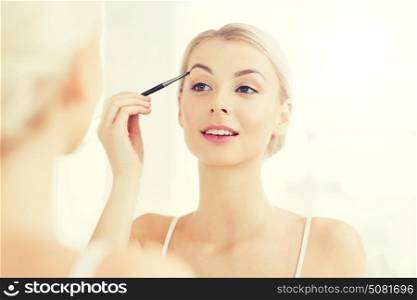 beauty, make up, cosmetics, morning and people concept - young woman applying makeup drawing eyebrow with brush and looking to mirror at home bathroom. woman with brush doing eyebrow makeup at bathroom
