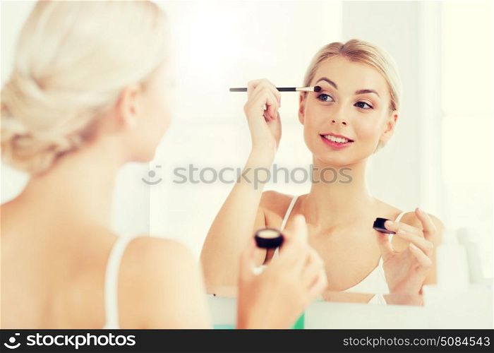 beauty, make up, cosmetics, morning and people concept - young woman applying eyeshade with makeup brush and looking to mirror at home bathroom. woman with makeup brush and eyeshade at bathroom. woman with makeup brush and eyeshade at bathroom