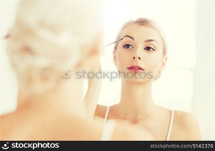 beauty, make up, cosmetics, morning and people concept - young woman applying makeup drawing eyebrow with brush and looking to mirror at home bathroom. woman with brush doing eyebrow makeup at bathroom