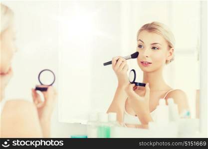 beauty, make up, cosmetics, morning and people concept - young woman applying blush with makeup brush and looking to mirror at home bathroom. woman with makeup brush and blush at bathroom